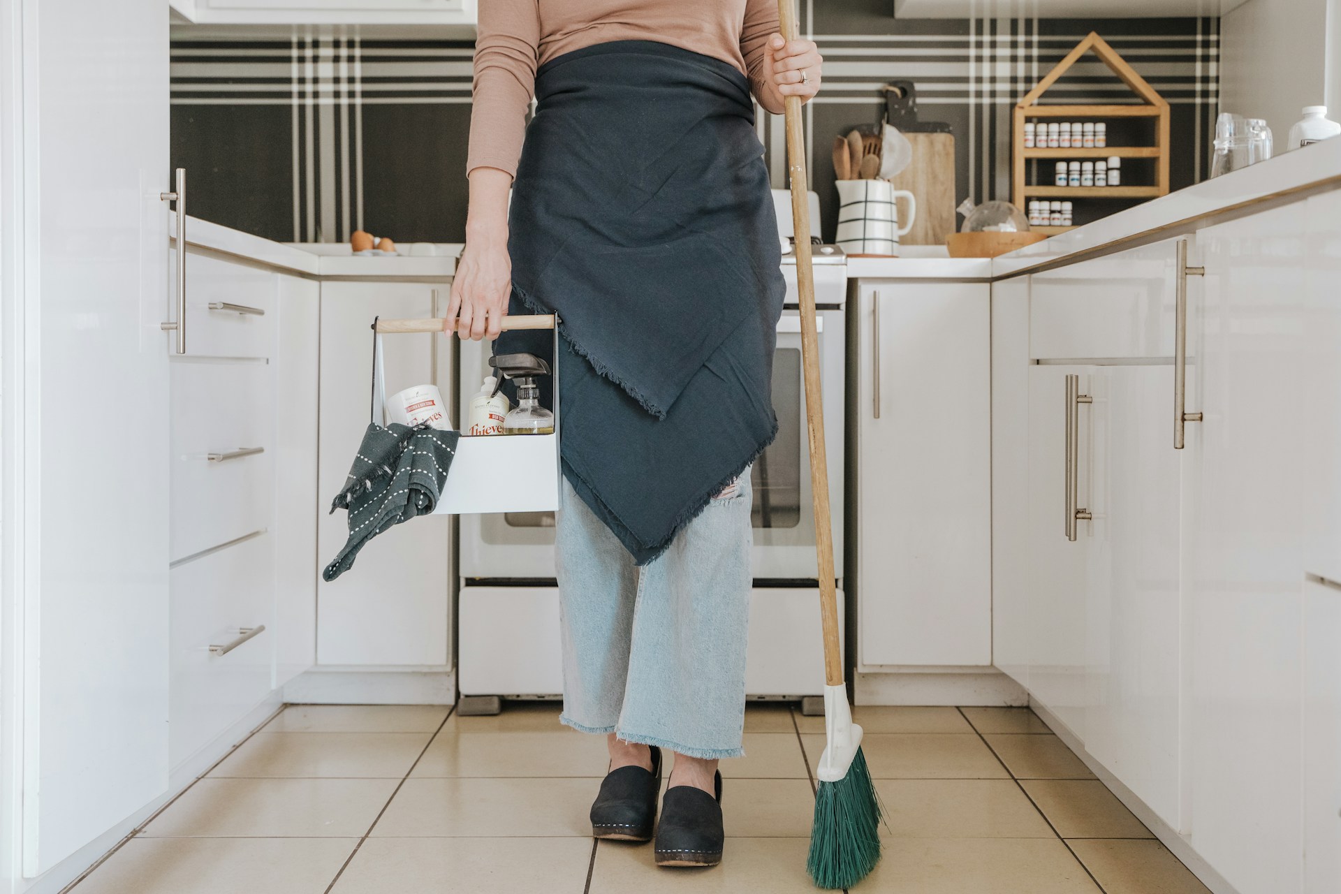 Cleaning professional holding a broom and caddy of supplies in a bright kitchen